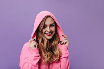 Lady in pink hoodie smiling on purple background. Portrait of blonde woman in sweatshirt looks...