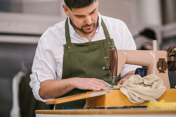 Diligent focused male tailor in apron sewing outfit details using modern sewing machine at table while creating exclusive clothes collection in contemporary work studio