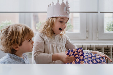 Excited charming girl in handmade crown unwrapping gift box while having birthday celebration at home