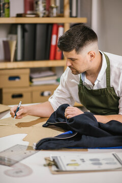Crop young male dressmaker writing with pen on sewing pattern examining or developing details of future outfit while working on exclusive apparel collection at table against blurred interior of contemporary atelier