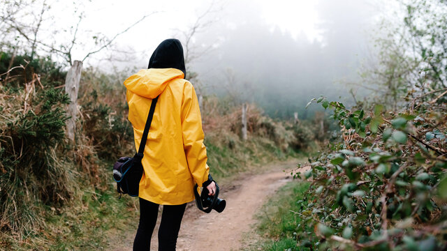 Back view of unrecognizable female tourist in vivid yellow raincoat and black hood with digital camera standing alone on road among foggy forest