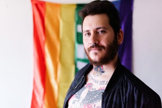 young Caucasian boy with a beard and tattoos poses in front of a rainbow flag of peace with a military jacket.