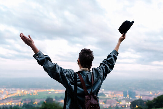 Back view of unrecognizable young lady in casual clothes with backpack and hat standing on hill with raised hands and admiring cityscape while spending time in evening