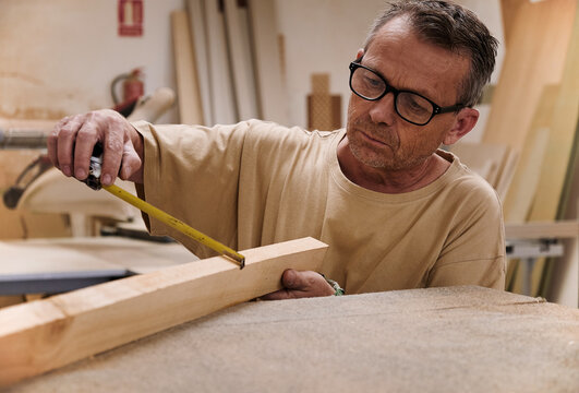 Crop focused carpentry worker in glasses and casual clothes controlling size of wooden detail using measuring tape while working in light modern workshop