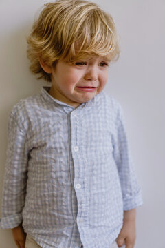 Disappointed little child in casual shirt standing near white wall and weeping on white background in studio