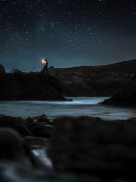 Silhouette of traveler standing with lantern on rocky shore of river and admiring mysterious scenery of tranquil water during starry night in highlands