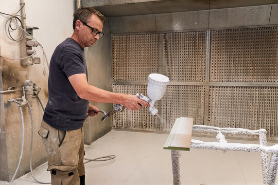 Side view of serious middle aged carpenter using spray gun for painting wood detail with white foamy flame retardant while working in light modern workroom