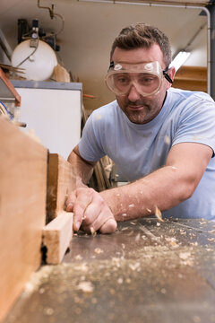 Low angle of focused carpenter in protective glasses milling timber while crafting detail using electric machine in light modern workshop