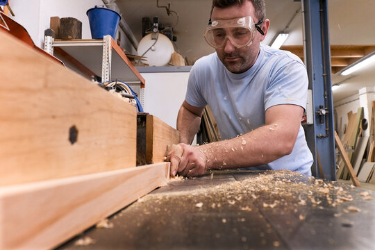 Low angle of focused carpenter in protective glasses milling timber while crafting detail using electric machine in light modern workshop