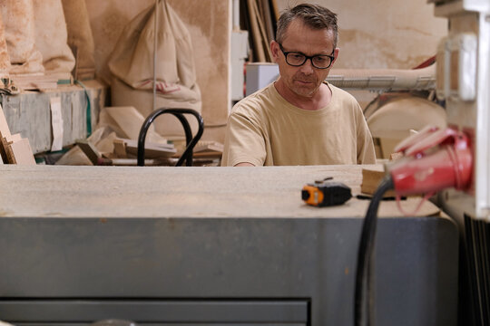Attentive middle aged workman in glasses and casual clothes focusing and using electric machine while working with timber in light modern carpentry studio