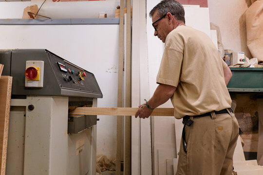 Low angle side view of crop attentive middle aged craftsman in glasses and casual clothes focusing and using carpentry electric machine while planing lumber in light modern studio