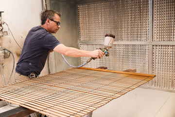 Adult focused man using spray gun painting wood slats with lacquer while working in contemporary carpentry workroom