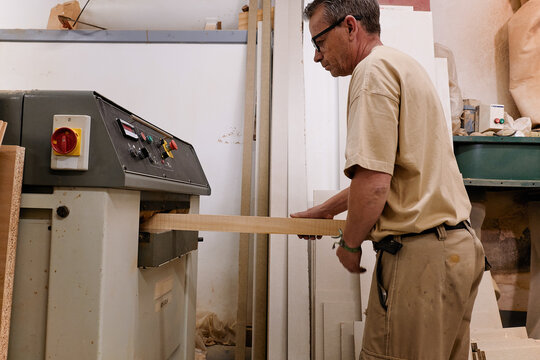 Low angle side view of crop attentive middle aged craftsman in glasses and casual clothes focusing and using carpentry electric machine while planing lumber in light modern studio
