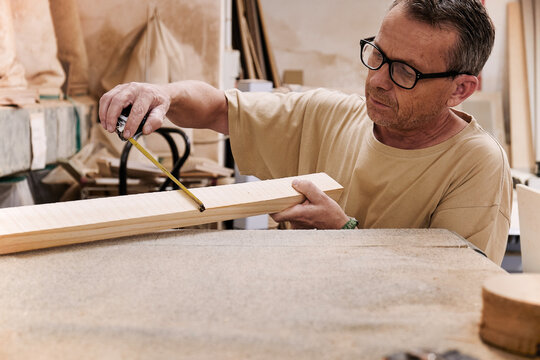 Crop focused carpentry worker in glasses and casual clothes controlling size of wooden detail using measuring tape while working in light modern workshop