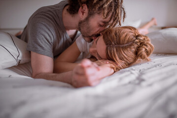 Tender man with dreadlocks hugging and kissing woman with red hair while lying on stomach on bed together and relaxing on weekend