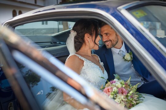 Happy bride in wedding dress and groom in classy suit sitting in vintage automobile and looking at each other