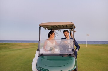 Smiling bride with bouquet of flowers and handsome groom in classy suit sitting in golf cart and driving across green lawn