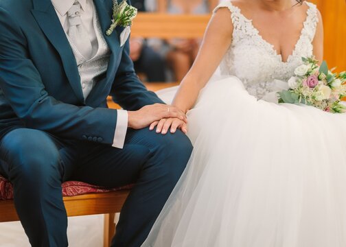 Unrecognizable groom in elegant suit with boutonniere and bride in white wedding gown sitting on bench and holding hands during ceremony in church