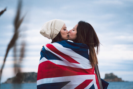 Side view of female lovers wrapping together in flag of Great Britain while hugging and kissing with closed eyes against sea coast and cloudy sky during romantic weekend