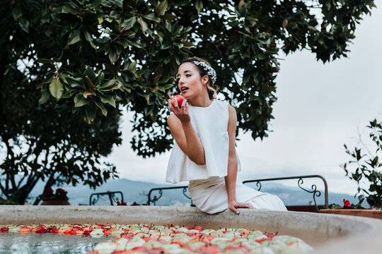 Elegant young woman sitting on fountain bowl and biting fresh apple on wedding day in garden
