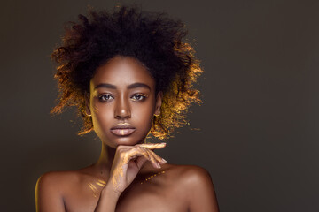 Portrait of a charming african girl with black curly hair and patterns of gold paint on her skin posing on a gray background in the studio