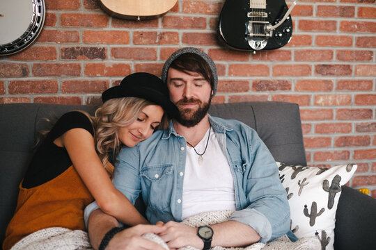 Smiling boyfriend in denim jacket embracing girlfriend in hat while sitting with closed eyes on cozy couch covered with rug blanket near brick wall with musical instruments