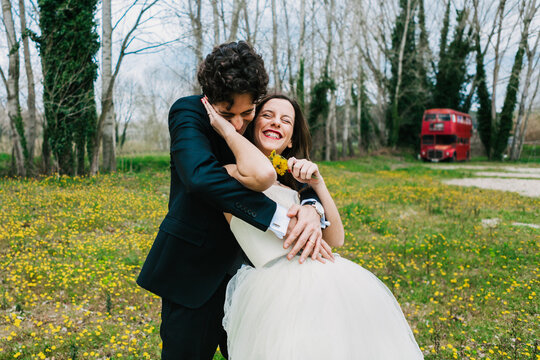 Happy groom in elegant suit hugging smiling bride with yellow dandelions on blooming meadow during wedding day