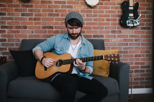 Cheerful bearded guitarist in sock cap and denim jacket playing acoustic guitar while sitting on couch with decorative pillow near brick wall with different musical instruments at home