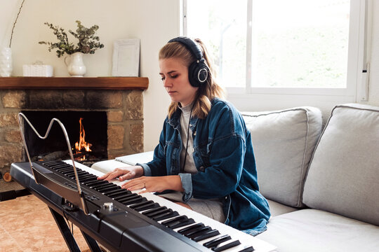 young female in casual wear sitting on sofa near fireplace and playing electric piano while spending time at home