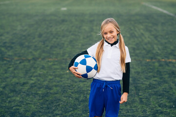 Cheerful preteen girl in white and blue uniform with soccer ball smiling at camera while standing alone on green field in modern sports club