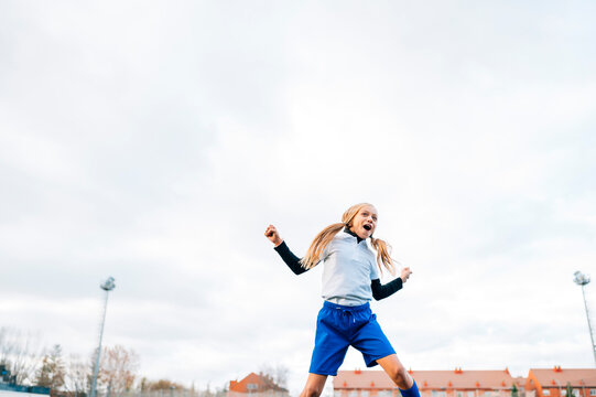 Excited teenage girl in white and blue uniform jumping with arm raised and screaming while celebrating victory in soccer match on green field in contemporary sports club