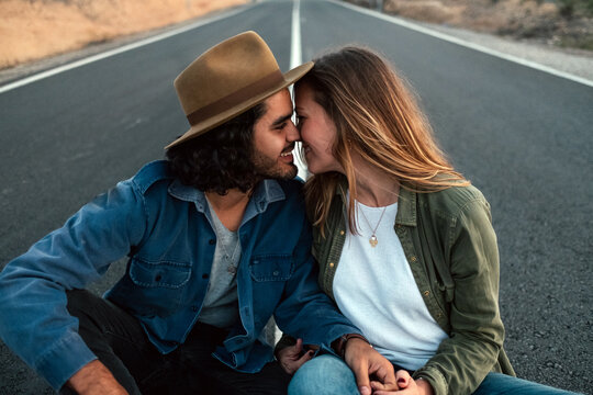 Cheerful man and woman in casual clothes smiling and touching noses while sitting on road in countryside