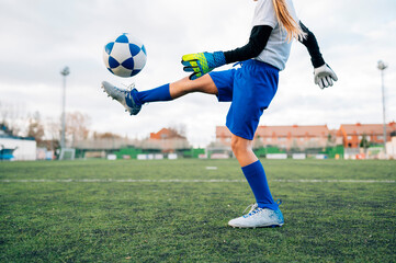 Low angle side view of unrecognizable teenage girl in white and blue uniform and goalkeeper gloves kicking ball while training alone in football arena at stadium in daytime