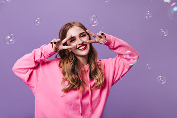 Happy lady in pink sweatshirt smiling and showing peace sign on purple background with bubbles. Joyful woman in hoodie laughing on isolated backdrop