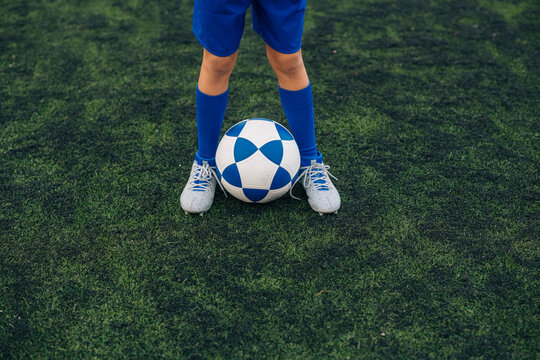 High angle of faceless child in blue shorts and cleats with ball standing alone on green court while playing football at modern stadium