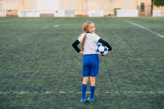 Girl in white and blue uniform with soccer ball smiling at camera while standing alone on green field in modern sports club
