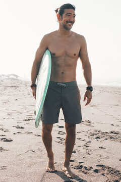 Happy shirtless ethnic male in shirts with blue surfboard looking away while standing on sandy beach near sea in sunny day