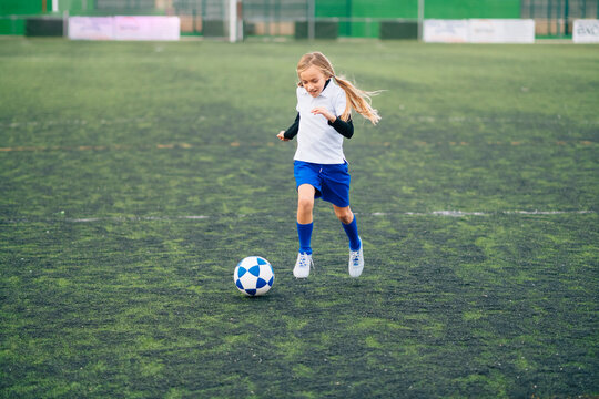 teenage girl in white and blue uniform and cleats running and preparing to kick ball while playing soccer alone on green field in contemporary sports club