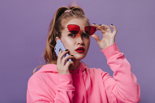 Portrait of girl in pink hoodie taking off red sunglasses and talking on phone. Closeup snaphot of young woman in street style sweatshirt posing on purple background