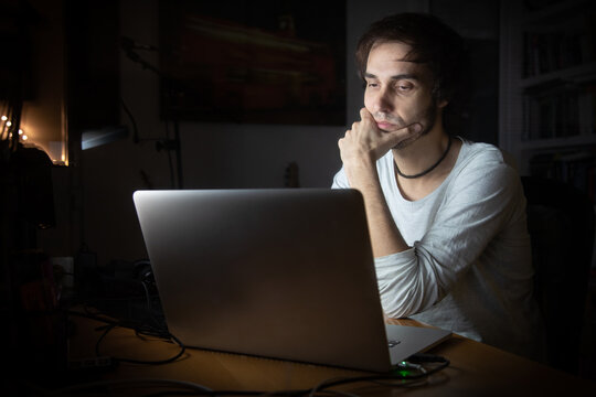 Concentrated young male remote specialist in casual wear talking on mobile phone and using laptop while working on project in dark room at home during evening time