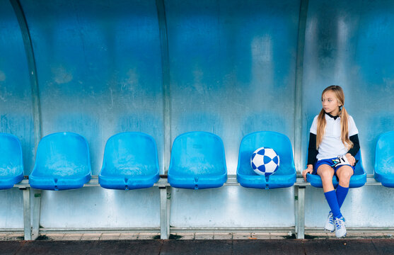 Full length frustrated preteen girl in soccer uniform sitting alone on blue plastic seat after match failure in sports club