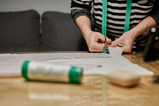 Cropped unrecognizable concentrated female designer in casual clothes standing near table and cutting fabric with scissors while working at home