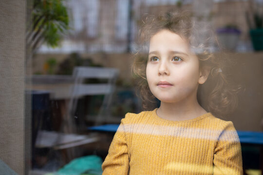 Calm little girl with curly hair standing near window and looking away thoughtfully while spending time at home and dreaming about adventures