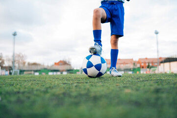 Low angle side view of unrecognizable teenage girl in white and blue uniform and goalkeeper gloves kicking ball while training alone in football arena at stadium in daytime