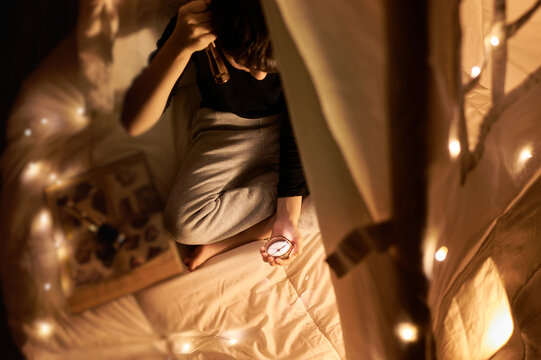 From above focused little kid in casual wear reading book while lying in kids play tent with flashlight and garland around near fireplace in apartment