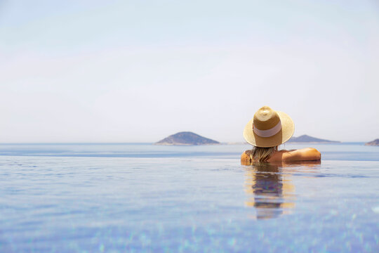 Young attractive woman enjoying the scenic ocean and mountain view from the edge of infinity pool on private villa. Back view of a sporty female in an outdoor swimming pool. Copy space, background.