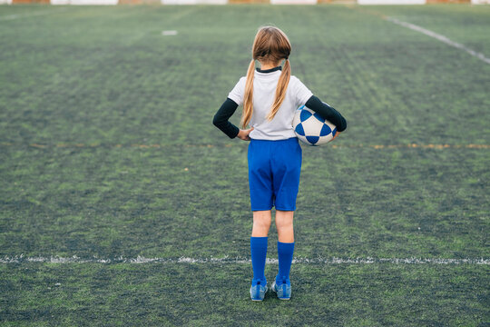 Girl in white and blue uniform with soccer ball smiling at camera while standing alone on green field in modern sports club