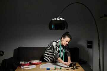 Focused female designer in casual clothes standing near table with tools and fabric while using smartphone to watch a tutorial during work at home