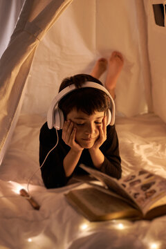 Thoughtful male child in headset reading book with images while listening to music and lying in cozy kids play tent in apartment