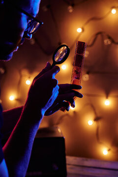 Side view of serious bald male photographer in casual clothes looking through magnifier while working with film in dark studio